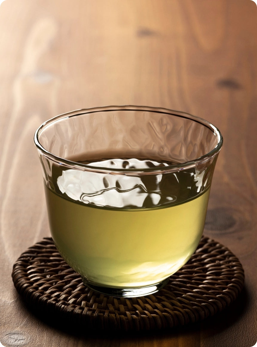 A clear glass cup filled with pale yellow tea, resting on a round woven dark brown coaster over a wooden table.