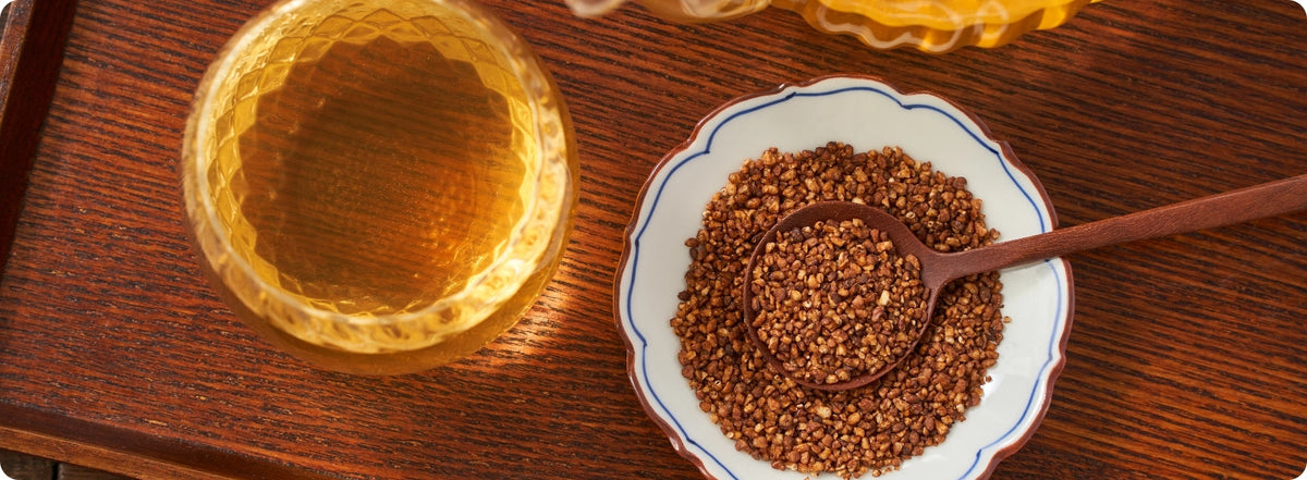 A glass of golden amber liquid beside a small white dish filled with brown seeds or grains, with a wooden spoon resting in the dish, all set on a dark wooden surface.