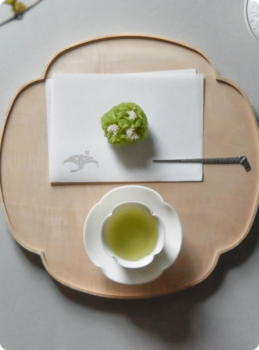A styled overhead shot of a light wood tray holding a white teacup with pale green tea and a small green flower-shaped pastry on a white napkin, with a metal spoon resting beside it.